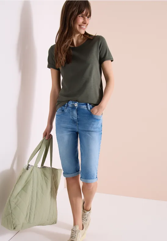 Frau in grünem T-Shirt, knielangen Jeans und Sneakers mit großer grüner Tasche, lächelnd.