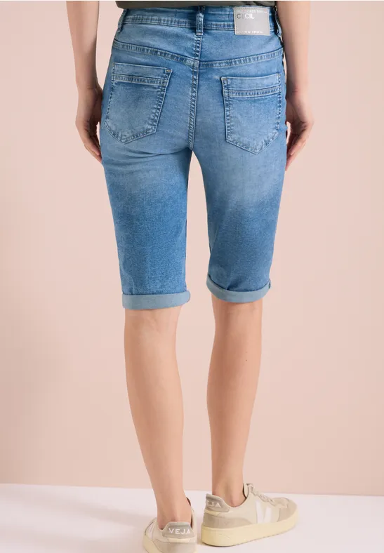 Hinteransicht einer Person in hellblauen Jeans-Bermudas bis übers Knie und Sneakers vor neutralem Hintergrund.
