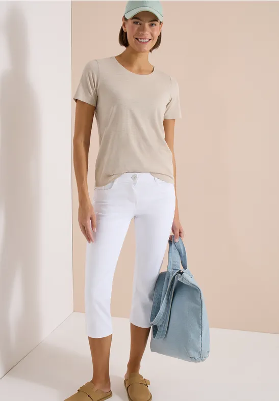 Frau in beigem T-Shirt, weißen Capri-Hosen, hält eine hellblaue Jeansjacke vor hellem Hintergrund.