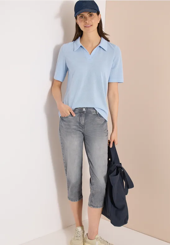 Frau trägt hellblauen Kurzarm-Pullover, graue Capri-Jeans und weiße Schuhe, hält eine blaue Jacke.
