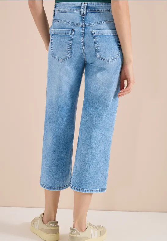 Rückansicht einer Frau in weit geschnittenen, knöchellangen Jeans und hellen Sneakern vor beigem Hintergrund.