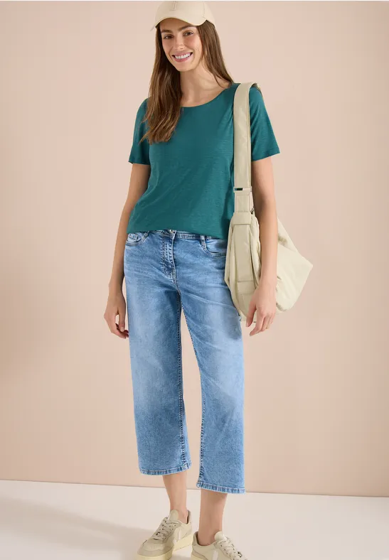 Frau trägt grünes T-Shirt, weite blaue Jeans und beigefarbene Sneaker, mit Stofftasche über der Schulter.