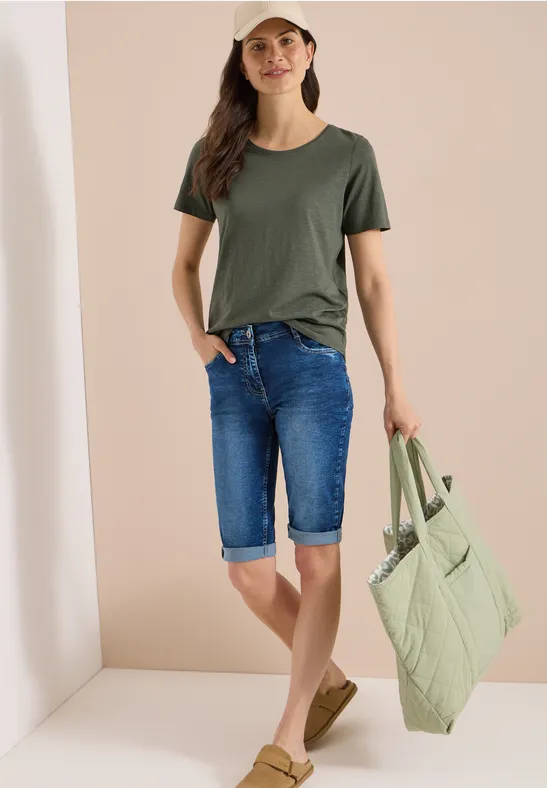 Frau in olivgrünem T-Shirt, Denim-Capri und hellgrünem gestepptem Shopper vor hellem Hintergrund.