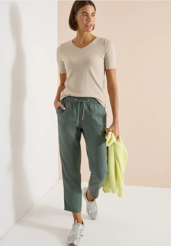 Femme en tee-shirt clair, pantalon vert à cordon, baskets, tenant une veste vert flashy.