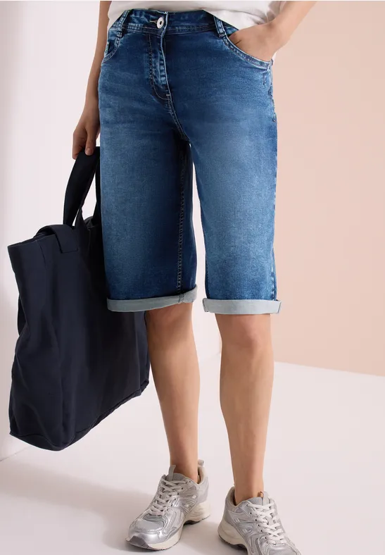 Une personne porte un short en jean bleu foncé retroussé, des baskets blanches et tient un tote bag noir.