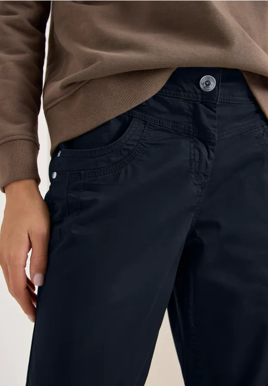 Nahaufnahme einer Person in dunklen Hosen und braunem Pullover mit Hand an der Tasche.