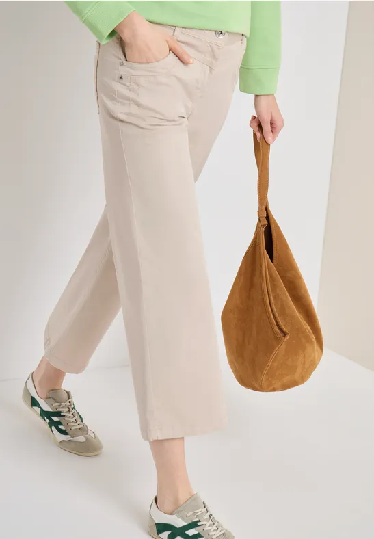 Modell trägt beigefarbene Karottenhose, weiße Sneaker mit grünen Details und braune Velours-Ledertasche