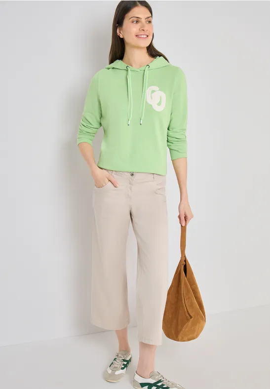 Person in grünem Hoodie, weiten Cremehosen, grünen Loafers und brauner Tasche vor hellem Hintergrund.