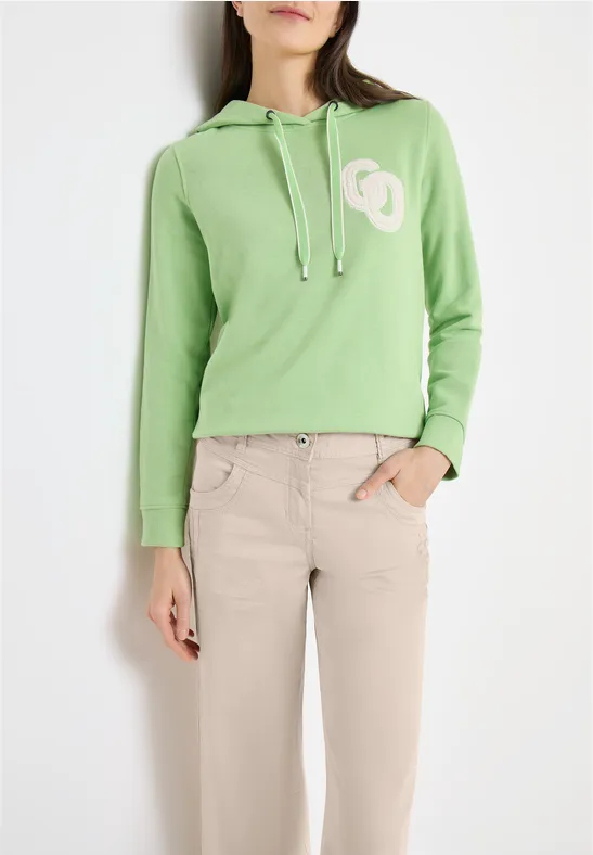 Frau trägt grünen Hoodie mit weißem Logo und beigefarbene Hose, lässiges Outfit.