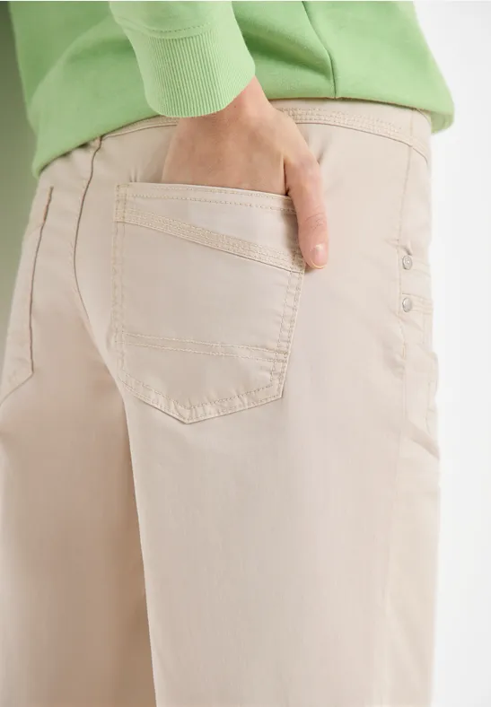 Person im hellgrünen Pullover steckt Hand in die Gesäßtasche einer hellbeigen Hose mit Knopfdetails.