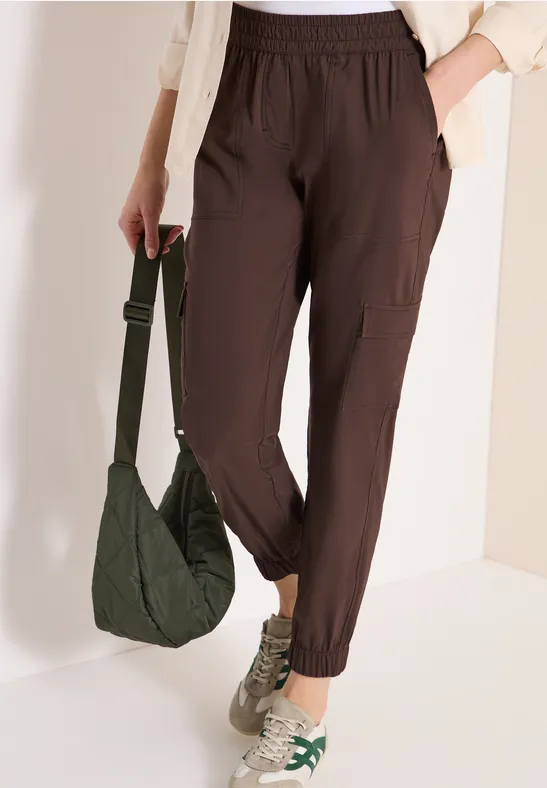 Eine Person trägt braune Cargo-Jogginghose, cremefarbene Sneaker und hält eine grüne Umhängetasche.