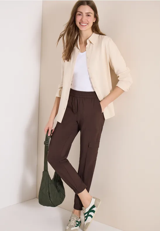 Frau trägt cremefarbene Strickjacke, weißes Top, braune Hose, weiße Sneaker und grünen Beutel.