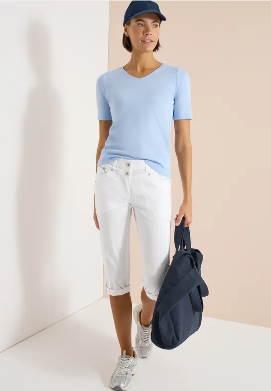 Frau in hellblauem T-Shirt, weißen knielangen Jeans und Sneakers mit dunkelblauem Rucksack.