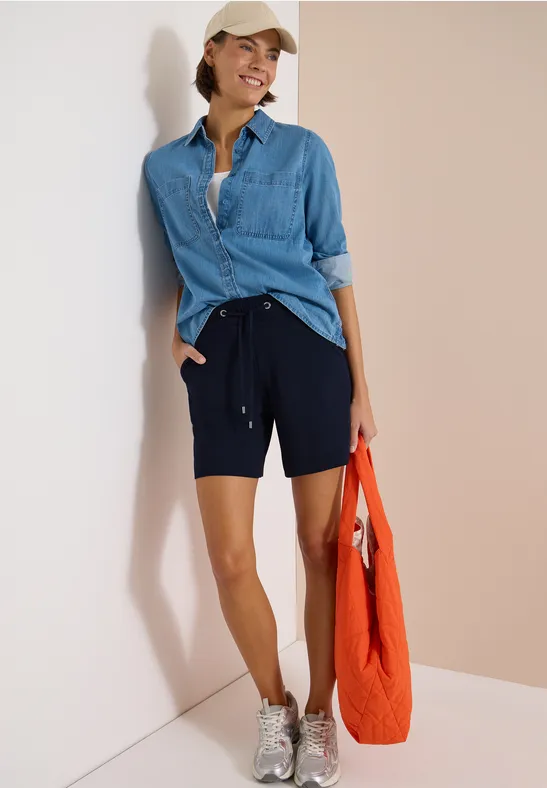 Eine Person lehnt an einer Wand, trägt Jeanshemd, schwarze Shorts, weiße Sneakers und hält eine orangefarbene Tasche.
