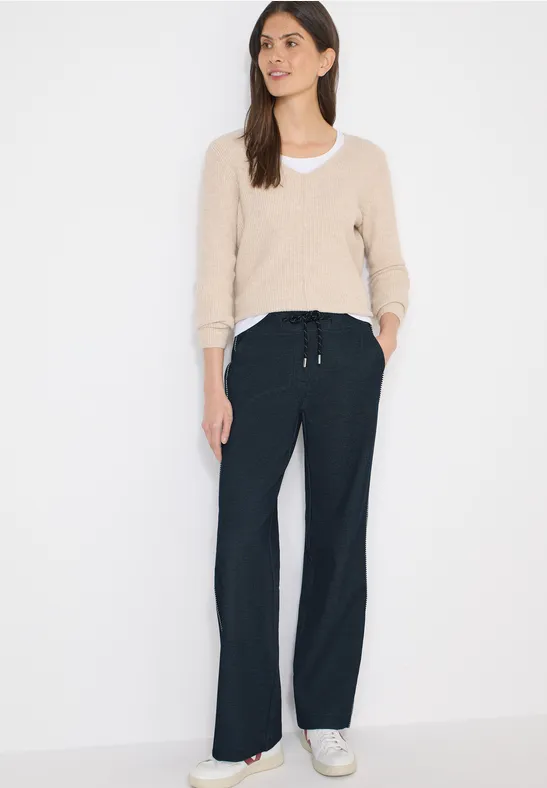 Person in beigem Pullover, schwarzer weiter Hose und weißen Sneakers vor weißem Hintergrund.