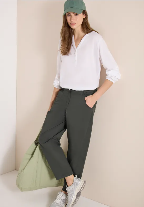 Person in weißer Bluse, dunkelgrauen Hosen und weißen Sneakers, hält eine grüne Tasche.