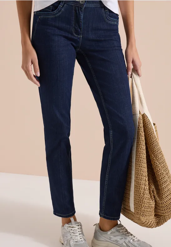 Eine Person trägt dunkle Jeans, weiße Sneaker und hält eine Strohtasche.
