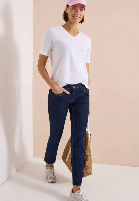 Frau trägt weißes V-Neck-T-Shirt, dunkelblaue Jeans, weiße Sneaker und geflochtene Tasche vor neutralem Hintergrund.