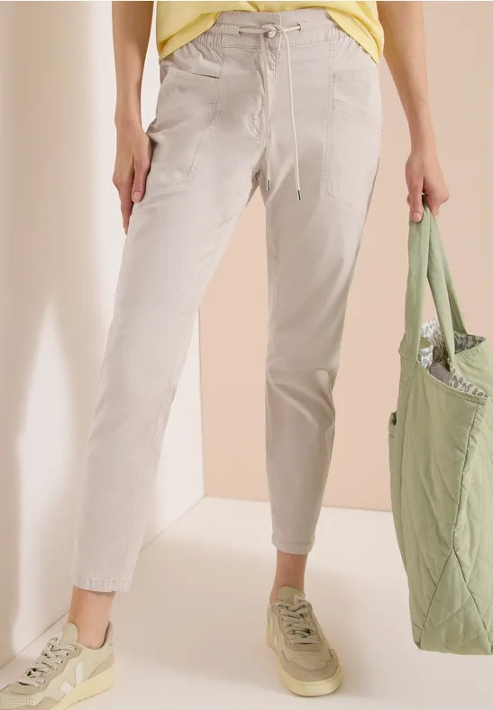 Eine Person trägt helle Stoffhosen, hellbeige Sneaker und hält einen grünen Stoffbeutel.