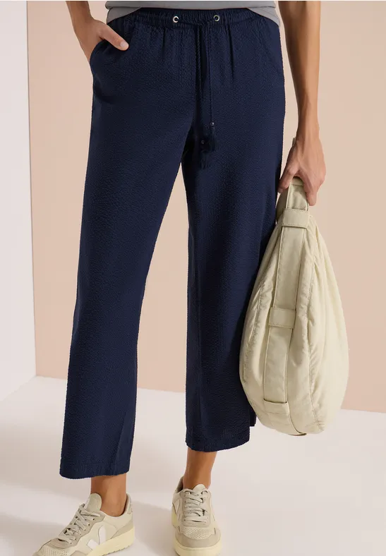 Marineblaue, locker sitzende Stoffhose mit Kordelzug, kombiniert mit hellen Sneakern und einer hellen Jacke in der Hand.