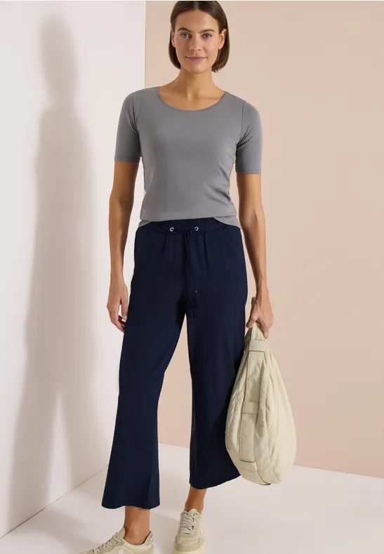 Frau mit grauem Shirt, weiten dunkelblauen Hosen und Tasche vor beigem Hintergrund.