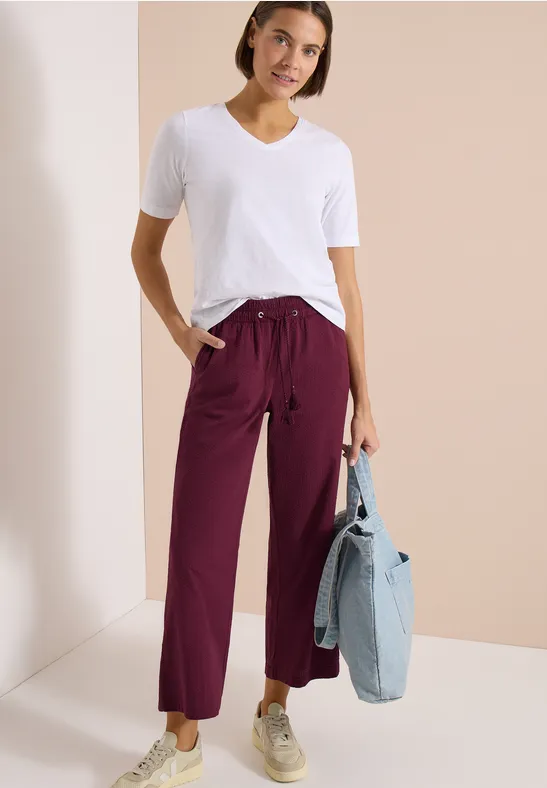 Frau trägt weißes T-Shirt, bordeauxrote Hose mit weitem Bein, beigen Sneakern und hält eine blaue Jeansjacke.