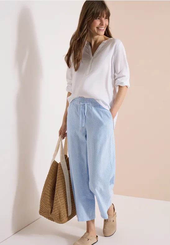 Frau in weißer Bluse und hellblauen, weiten, knöchellangen Jeans, mit Strohbeutel und Espadrilles.