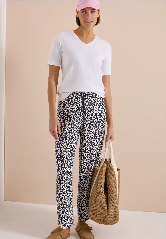 Frau trägt weißes T-Shirt, gemusterte Hose und hält großen Strohsack vor hellem Hintergrund.