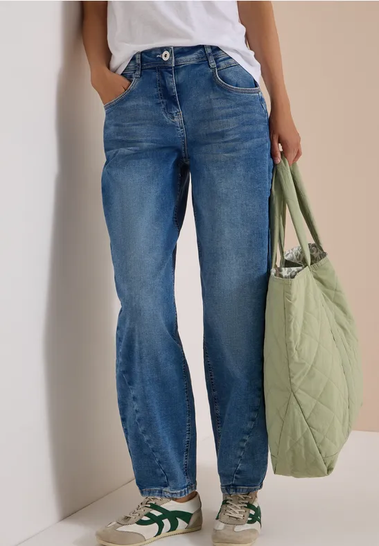 Person trägt blaue Jeans, weißes Top und hält eine große grüne Tasche.