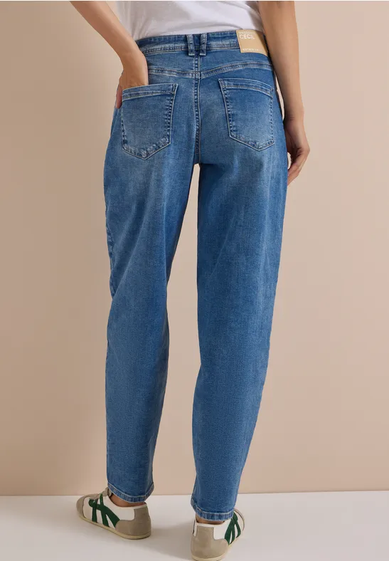 Person von hinten in blauen Jeans und Turnschuhen vor neutralem Hintergrund.