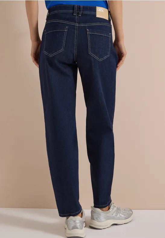 Eine Person steht von hinten, trägt dunkelblaue Jeans und helle Sneaker vor beigem Hintergrund.