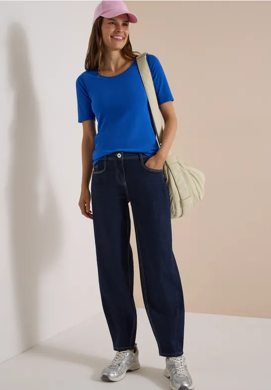 Frau mit rosa Cap, blauem Shirt, weiten Jeans, silbernen Sneakern und Tasche vor beigem Hintergrund.
