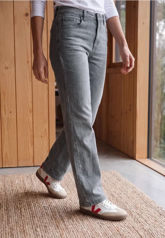 Jambes en jean gris clair et sneakers blanches à V rouge, posées sur un tapis près d'une fenêtre.