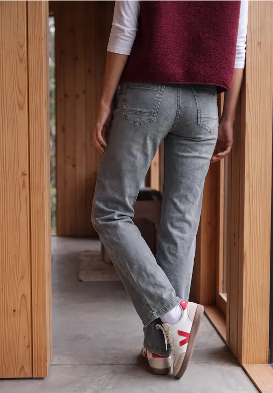Personne vue de dos en jean gris et baskets blanches, debout dans une ouverture en bois.