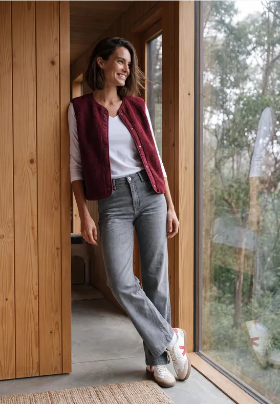 Femme en jean gris, t-shirt blanc et gilet rouge, appuyée à l'encadrement en bois près d'une fenêtre.