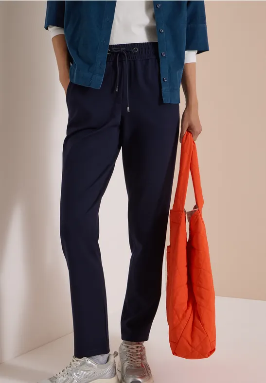 Person trägt dunkelblaue Freizeithose, weiße Sneaker und hält eine orangefarbene Tasche.