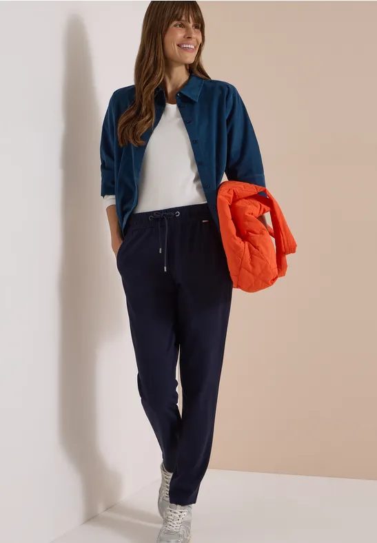 Frau lehnt an Wand, trägt weißes Shirt, blaues Hemd, dunkelblaue Hose und hält eine orange Jacke.