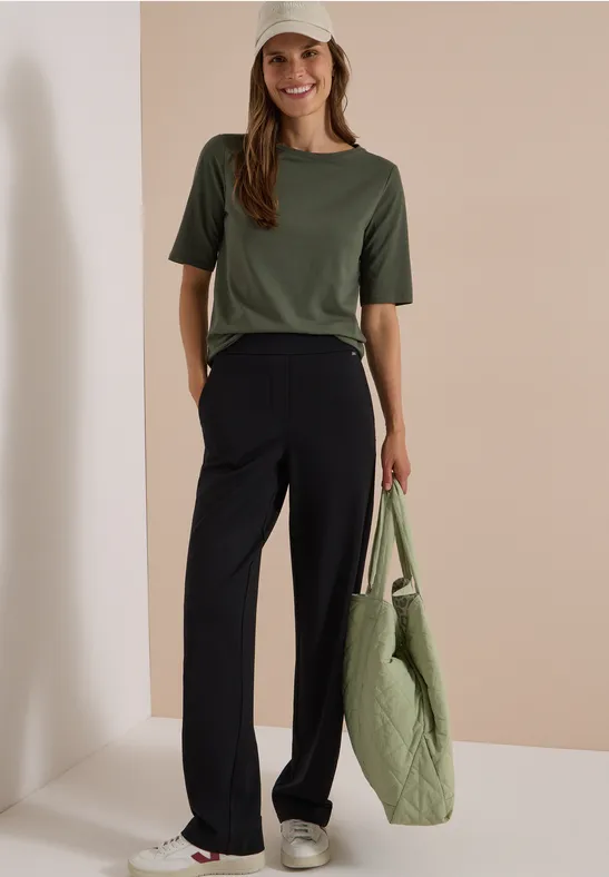Femme souriante en t-shirt kaki, pantalon noir ample et casquette, tenant un grand sac matelassé devant un mur beige.