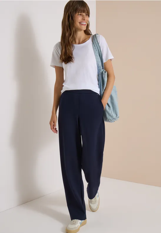 Femme souriante en t-shirt blanc et pantalon bleu marine ample, portant un sac en tissu léger sur l'épaule.