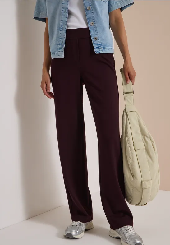 Frau trägt plumfarbene weite Hose, helle Jeansjacke, silberne Sneaker und hält einen beigen Stoffbeutel.