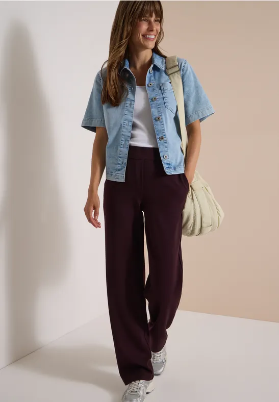 Frau in burgunderroter Marlene-Hose, weißem Top, jeansblauer Kurzjacke und Sneakers mit Tasche.