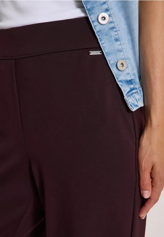 Nahaufnahme einer Person in weinroter Hose, weißem Oberteil und Jeansjacke mit Hand an der Seite