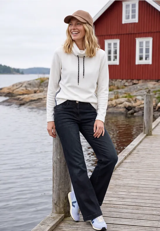 Femme en sweat blanc, jeans noirs évasés et baskets blanches sur un ponton devant une maison rouge au bord de l'eau.