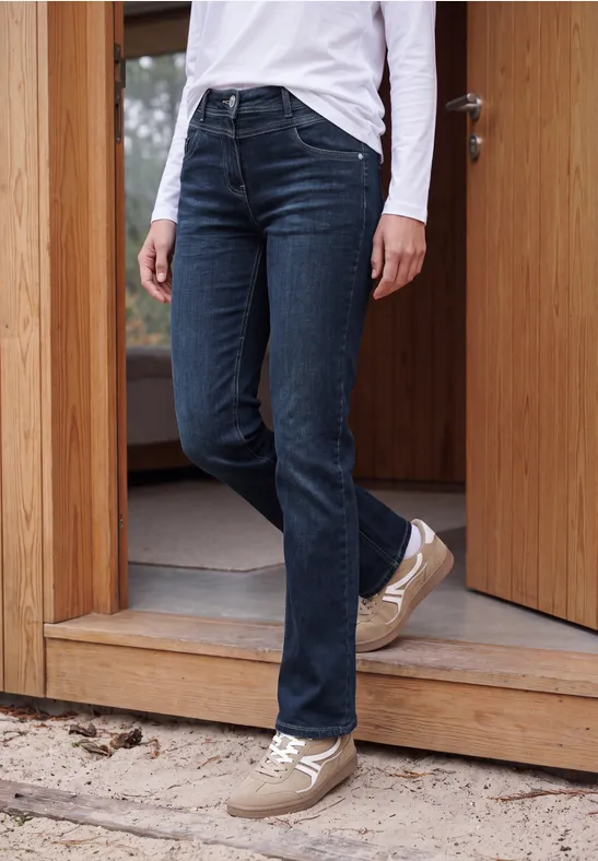 Personne en jean bleu foncé et baskets claires debout dans un cadre de porte en bois.