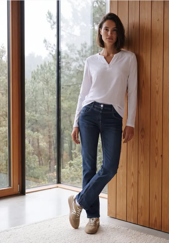 Une femme en jean et chemisier blanc pose détendue contre un mur de bois, face à une fenêtre donnant sur la forêt.