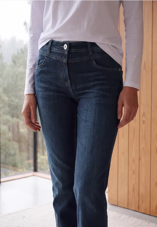 Une personne porte un jean bleu foncé et un haut blanc, debout près d’une fenêtre à côté d’un mur en bois.