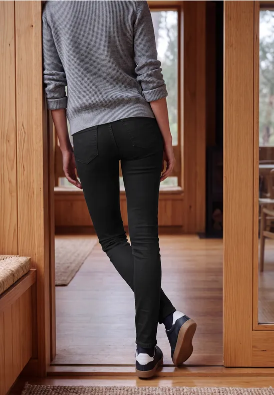 Vue arrière d’une personne en pantalon noir et pull gris, debout devant une porte en bois.