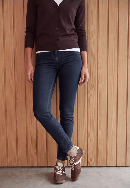 Vue partielle d'une personne en jean skinny foncé, baskets marron et gilet, adossée à un mur en bois.