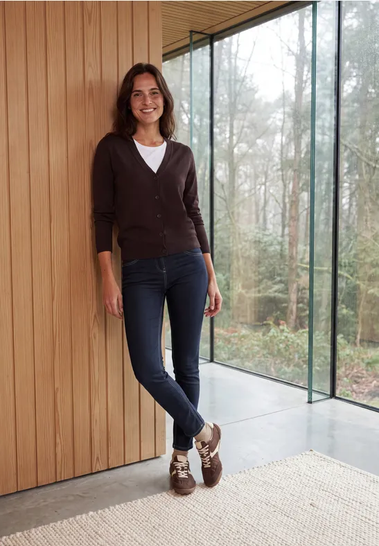 Femme souriante appuyée sur un mur en bois dans un intérieur moderne, devant une baie vitrée avec vue sur forêt.