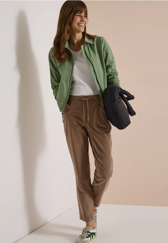 Une femme souriante adossée à un mur, veste verte, t-shirt blanc, pantalon marron et sac matelassé.